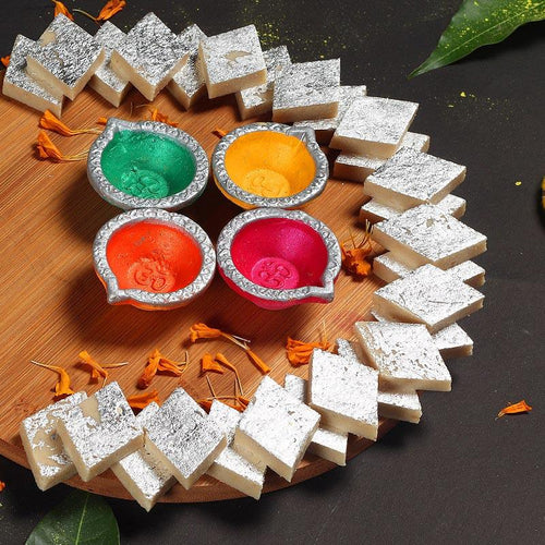 Decorative arrangement of white and silver Kaju Katli with colorful Diwali Diya on a wooden board.