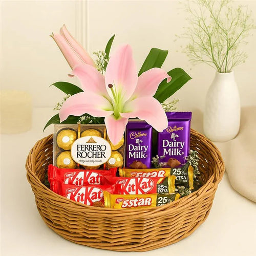 Basket of assorted chocolates and pink lilies on a light surface with a vase in the background.