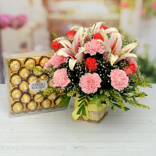 Floral arrangement with pink and red flowers and a box of Ferrero Rocher chocolates on a white surface.