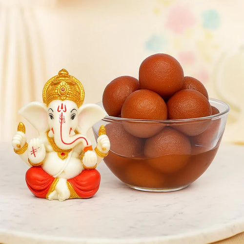 Small statue of Ganesha next to a glass bowl filled with round brown Gulab jamun on a light background.