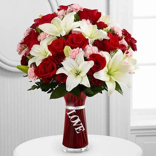 Mixed flower bouquet with red roses, white lilies, pink carnations in a glass vase labeled 'Love'
