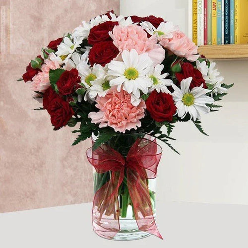Bouquet of red, white, and pink flowers in a clear vase with a red ribbon on a light background.