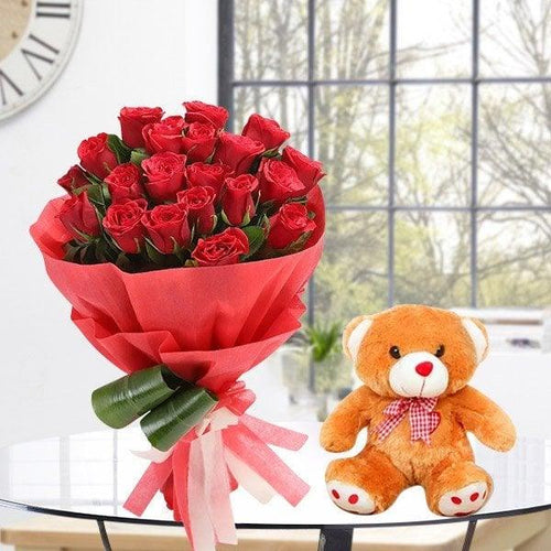 Bouquet of red roses wrapped in red paper with a teddy bear on a table.