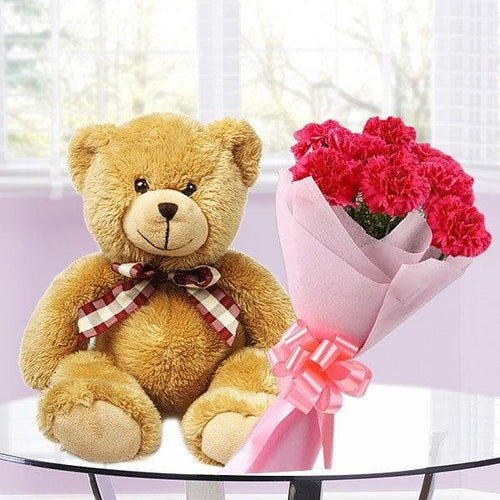 Teddy bear with a bow tie next to a bouquet of pink flowers on a white surface.