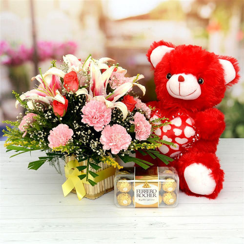 Red teddy bear, bouquet of flowers, and Ferrero Rocher chocolates on a table.