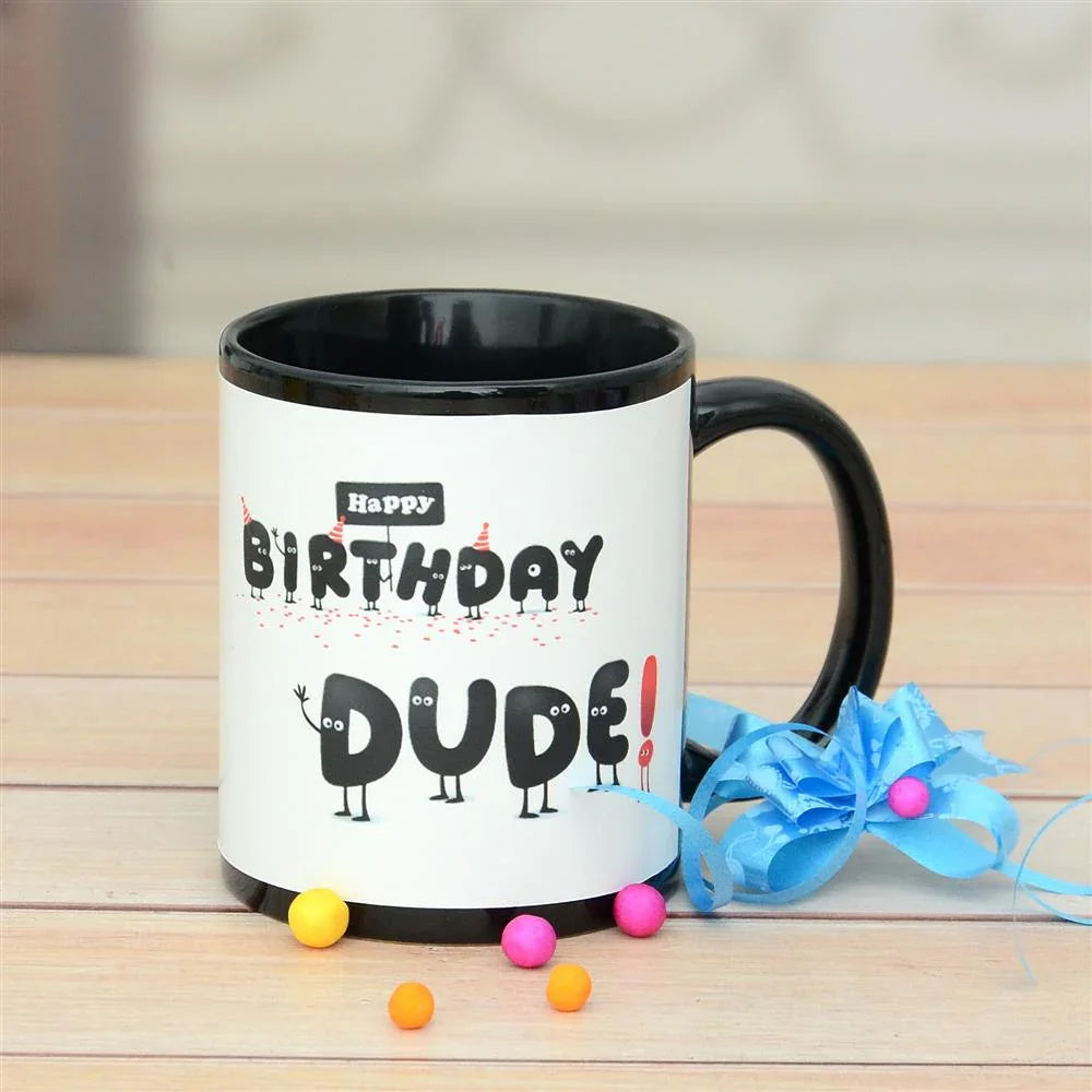 Black and white mug with 'Happy Birthday DUDE!' text on a wooden surface with colorful candies and a blue bow.