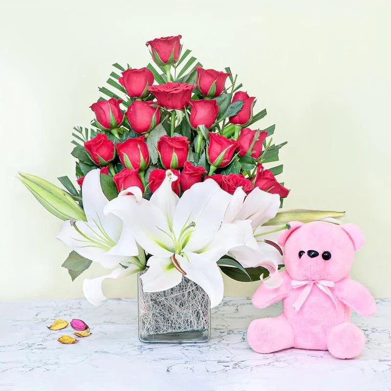 Bouquet of red roses and white lilies with a pink teddy bear on a light surface.