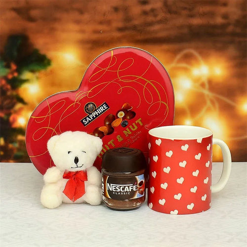 Heart-shaped chocolate box, teddy bear, Nescafe jar, and red mug with heart patterns on a blurred background.