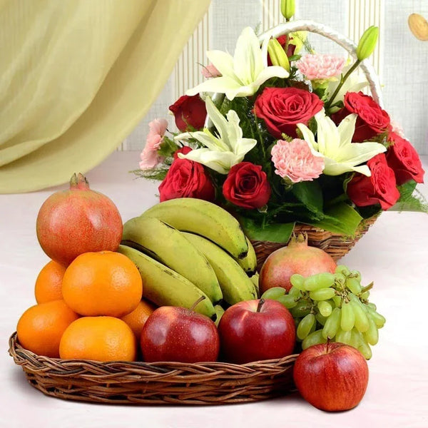 Basket of fruits including apples, oranges, and bananas, with a bouquet of flowers in the background.