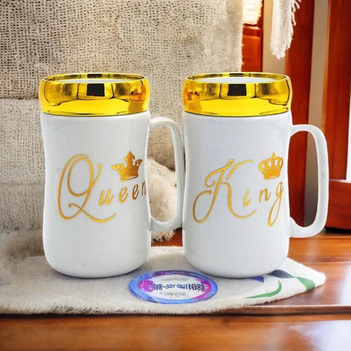 Two white mugs with gold lids, one labeled 'Queen' and the other 'King', on a wooden surface.