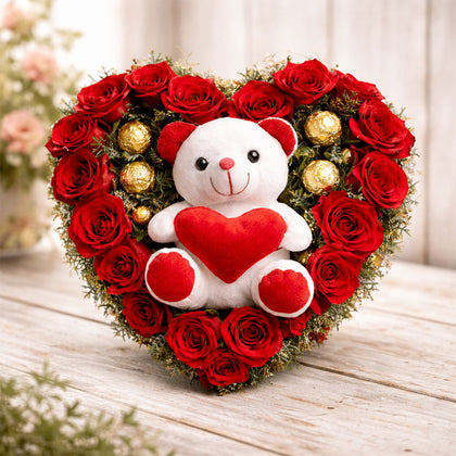 Heart-shaped arrangement of red roses with a teddy bear holding a red heart on a wooden surface.