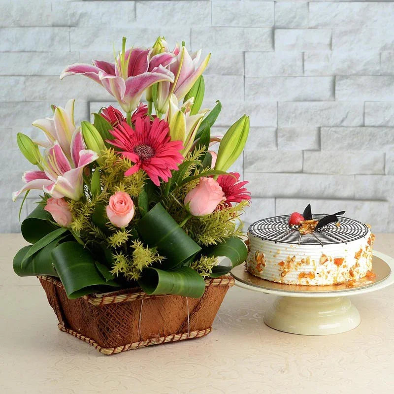 Floral arrangement in a basket next to a cake on a table with a light gray tiled wall background.