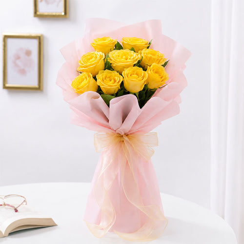 A bouquet of yellow roses wrapped in light pink tissue paper and a sheer pink ribbon, displayed on a white table against a softly lit indoor background.