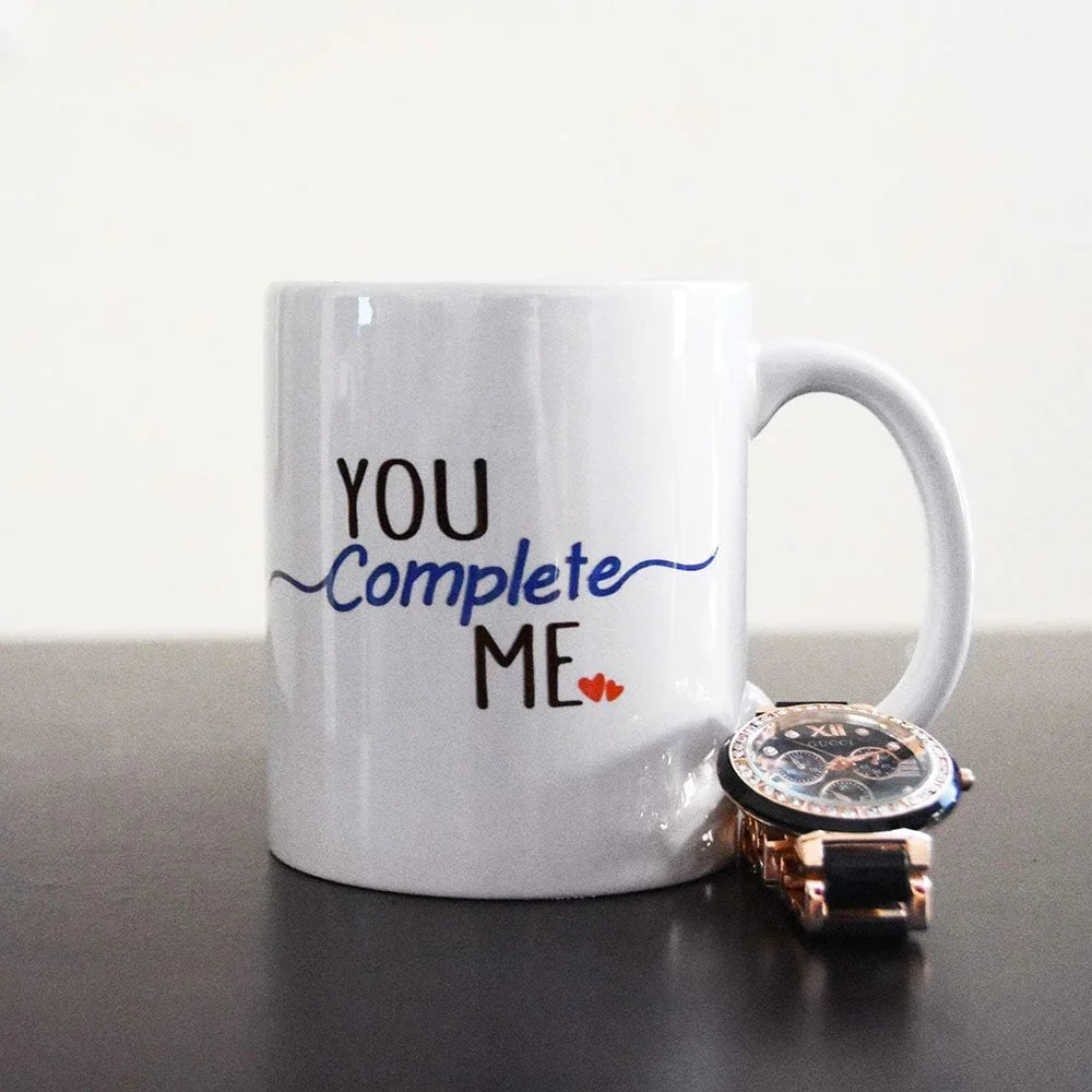 White mug with 'You Complete Me' text and a watch on a reflective surface.