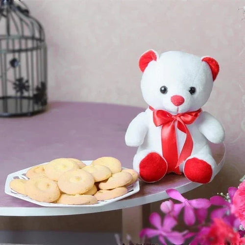 White teddy bear with a red bow tie sitting next to a plate of cookies on a table.