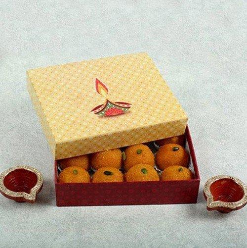 Box of Indian sweets with decorative lid, surrounded by two festive diyas on a light background