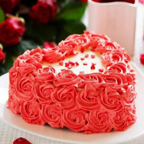 Heart-shaped cake with red rose decorations on a white plate, surrounded by flowers.