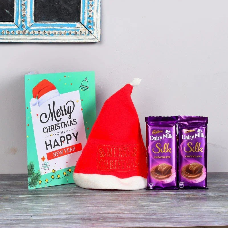 Red Santa hat, Christmas card, and Cadbury Dairy Milk Silk chocolate bars on a wooden surface.