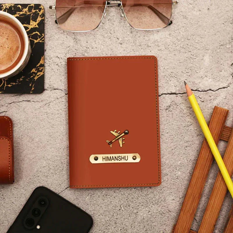 Brown leather passport holder with airplane emblem and 'HIMANSHU' engraving on a concrete surface with coffee, sunglasses, and pencils.