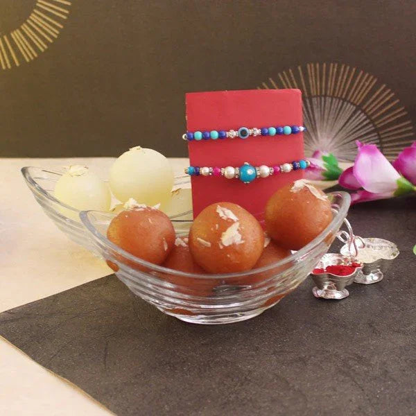 Glass bowl with gulab jamun and rasgulla sweets, colorful rakhi bracelets, flowers, and traditional puja items for Raksha Bandhan celebration.