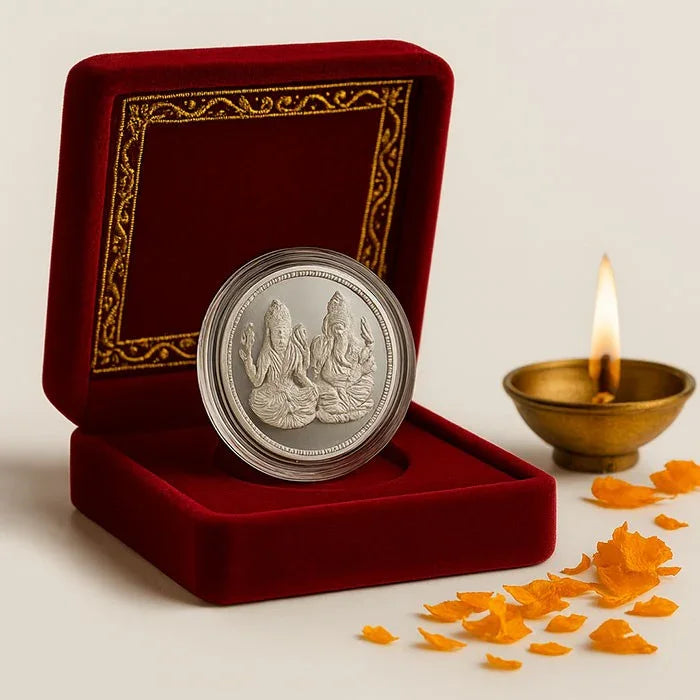 Silver coin with engravings in a red velvet box with gold detailing, next to a lit diya and flower petals.