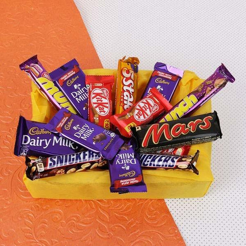 Assorted chocolate bars including Mars, Cadbury, and KitKat in a yellow basket on an orange and white background.