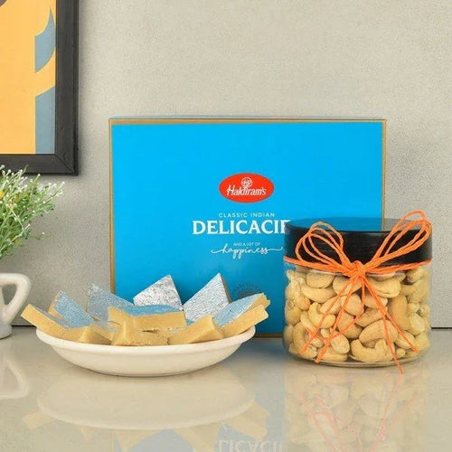 Plate of kaju katli sweets with silver leaf, jar of cashews, and blue Haldiram's gift box
