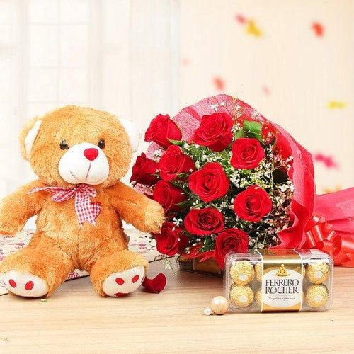 Teddy bear, bouquet of red roses, and Ferrero Rocher chocolates on a wooden surface.