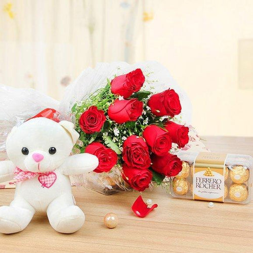 White teddy bear with a pink bow, bouquet of red roses, and Ferrero Rocher chocolates on a wooden surface.