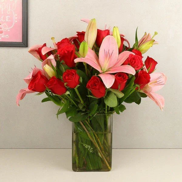 Bouquet of red roses and pink lilies in a clear vase against a light gray wall.