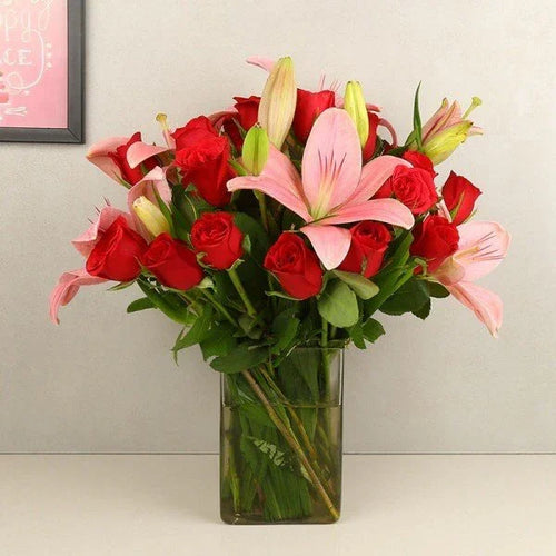 Bouquet of red roses and pink lilies in a clear vase against a light gray wall.