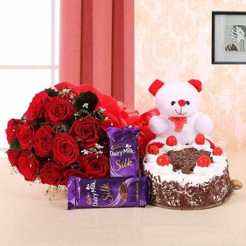 Bouquet of red roses, a teddy bear, a cake, and Cadbury Dairy Milk Silk chocolate bars on a table.