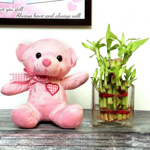 Pink teddy bear with a heart and a glass vase with green bamboo plant on a wooden surface.