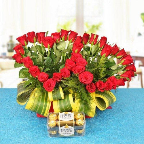 Heart-shaped arrangement of red roses with green leaves on a blue tablecloth, accompanied by Ferrero Rocher chocolates.