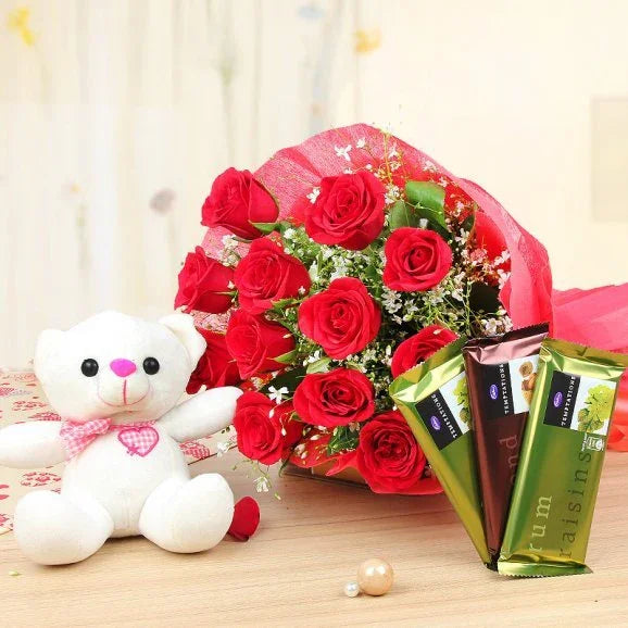 Bouquet of red roses with a white teddy bear and chocolate bars on a wooden surface.