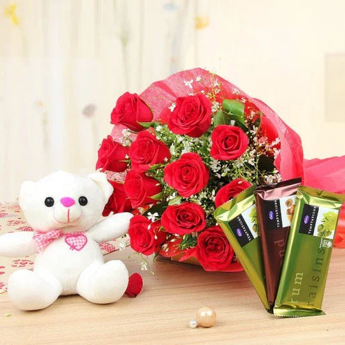 Bouquet of red roses with a white teddy bear and chocolate bars on a wooden surface.