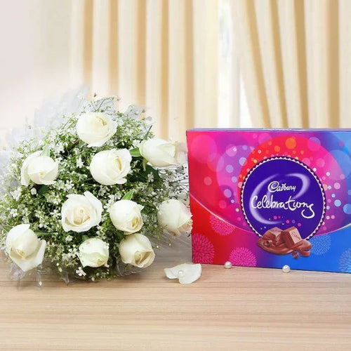 Bouquet of white roses and Cadbury Celebrations chocolate box on a wooden surface with beige curtains in the background.
