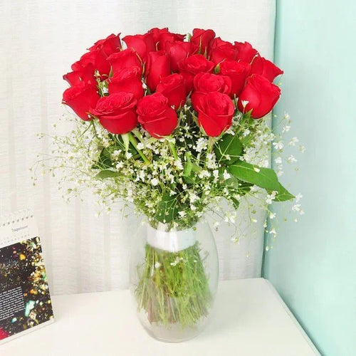 Bouquet of red roses with greenery in a clear vase on a white surface.